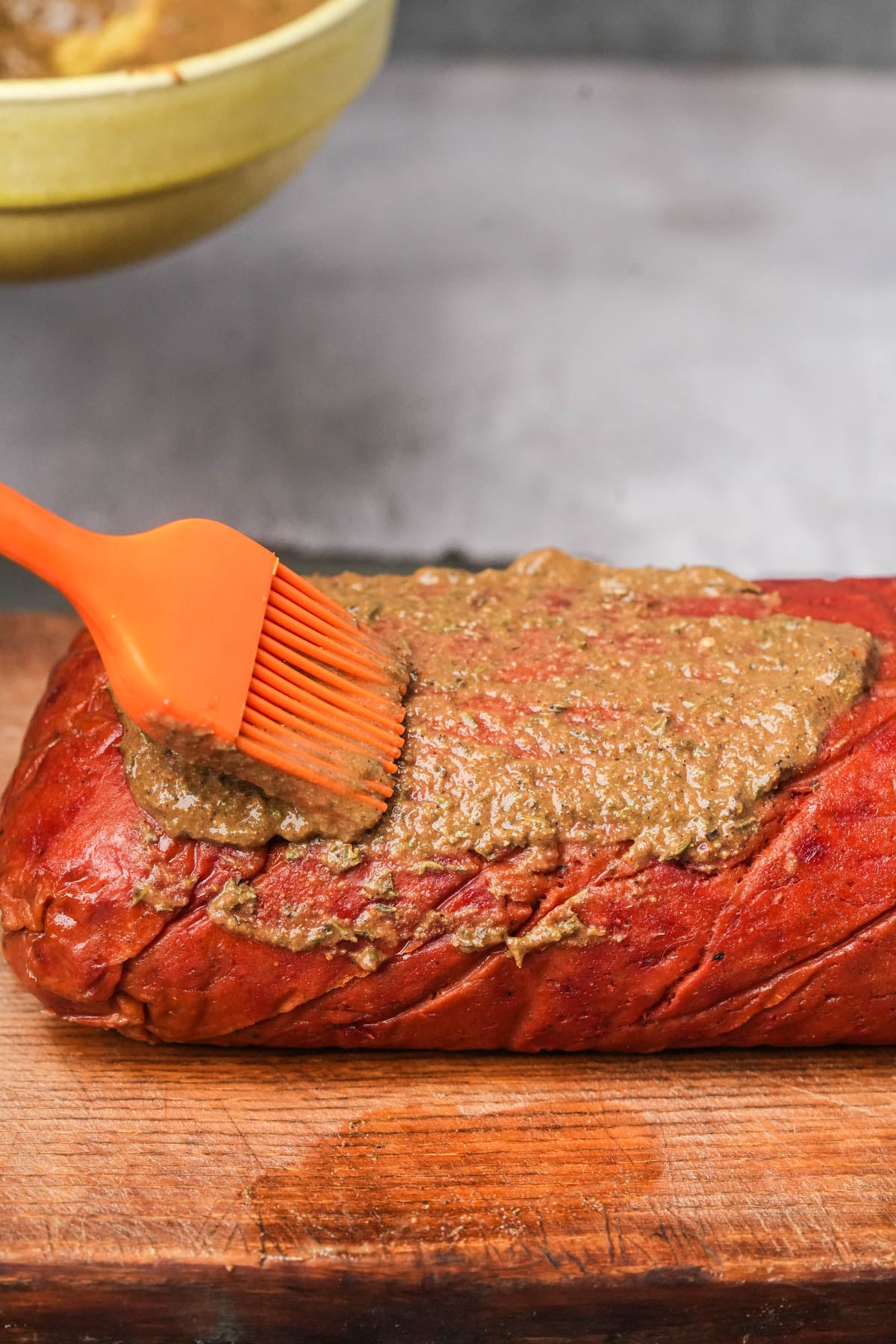 An orange brush spreads a thick, brown marinade on a piece of steamed vegan roast beef resting on a wooden cutting board. A yellow bowl with more marinade is visible in the background.