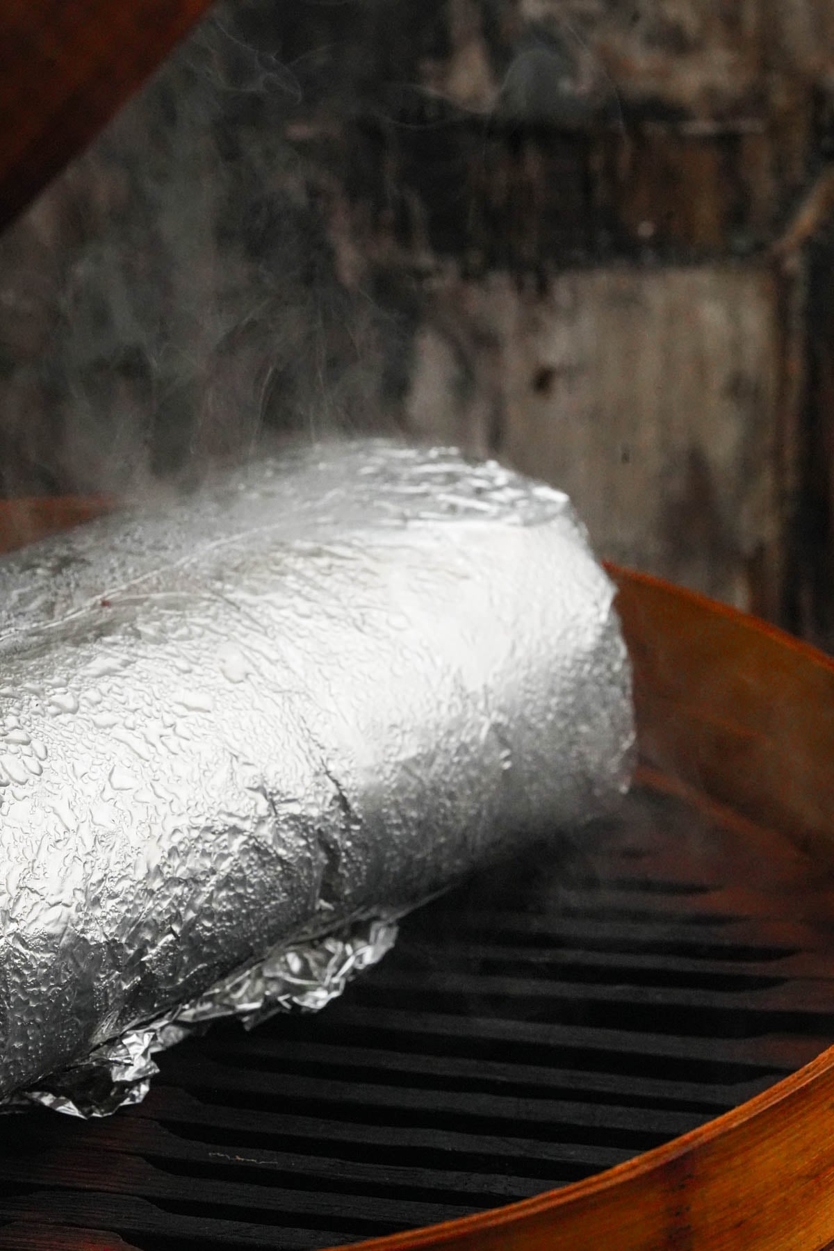 A large, foil-wrapped log of vegan roast beef sits on the grate of a steamer, with steam rising from it. The background is rustic and wooden.