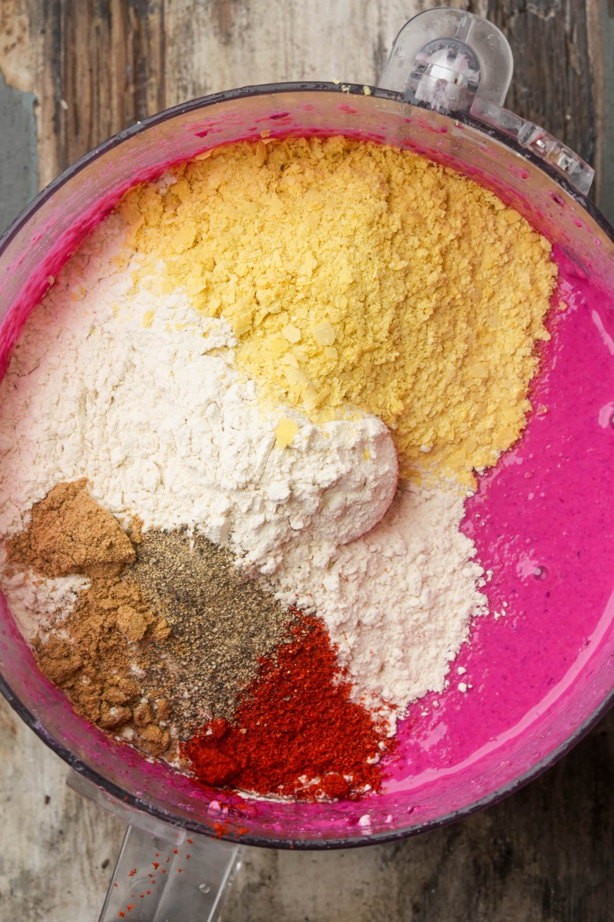 A food processor bowl filled with bright pink blended beet base, topped with piles of vital wheat gluten, nutritional yeast, spices and powders, ready to be mixed. The bowl sits on a rustic wooden surface.
