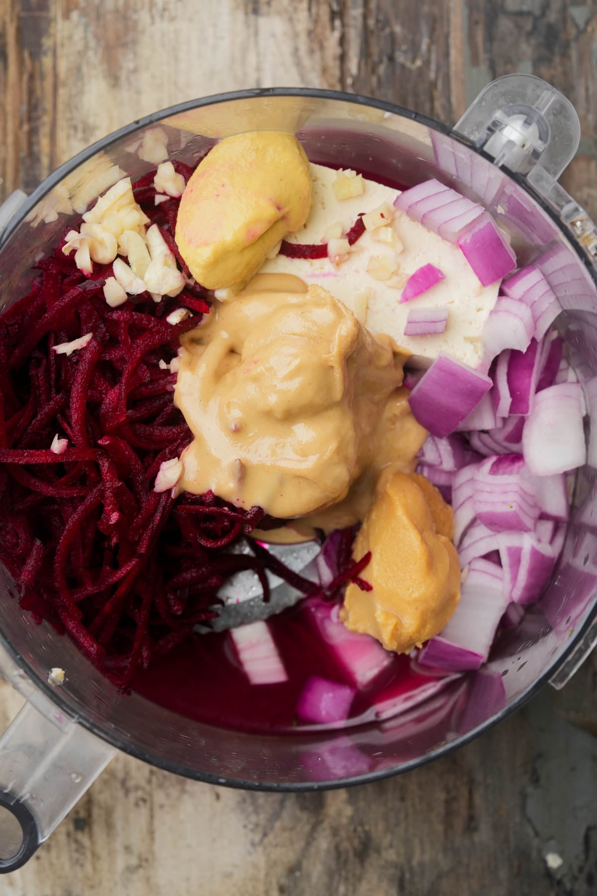 A food processor bowl filled with chopped red onion, grated beet, garlic, two scoops of tahini, and mustard, all sitting on a rustic wooden surface.
