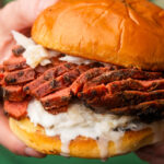 A close-up of a hand holding a sandwich with sliced Vegan Roast Beef, creamy white sauce, and shredded cabbage on a shiny, toasted bun.