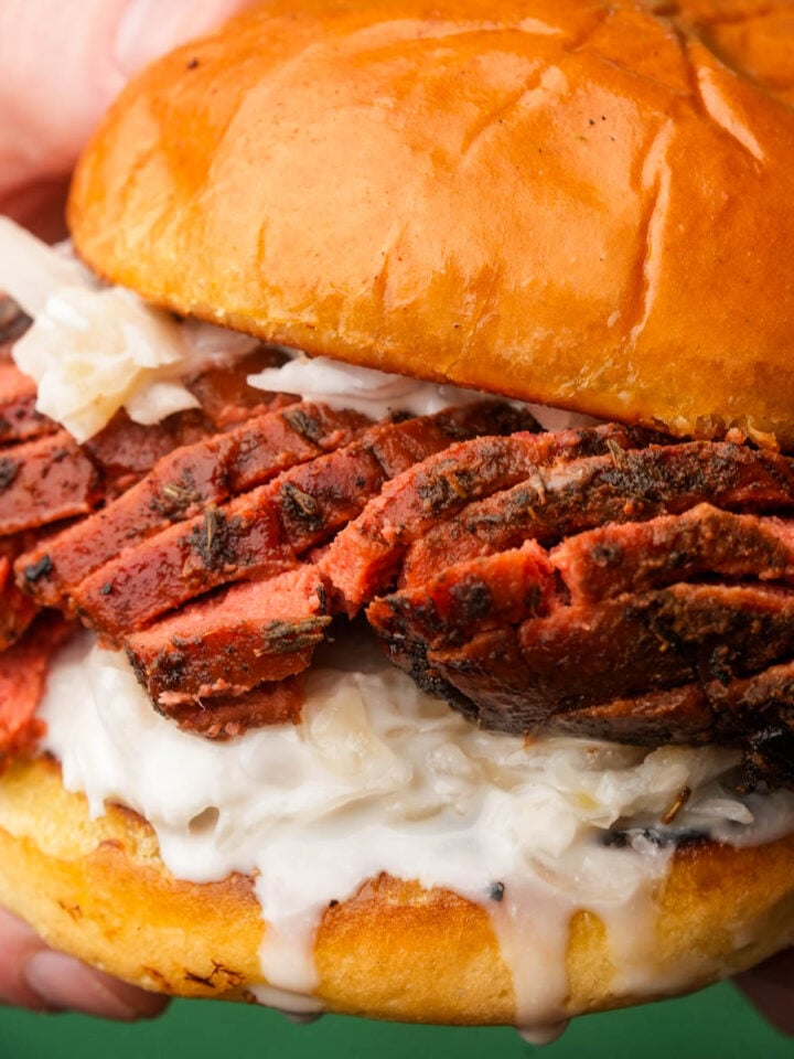 A close-up of a hand holding a sandwich with sliced Vegan Roast Beef, creamy white sauce, and shredded cabbage on a shiny, toasted bun.