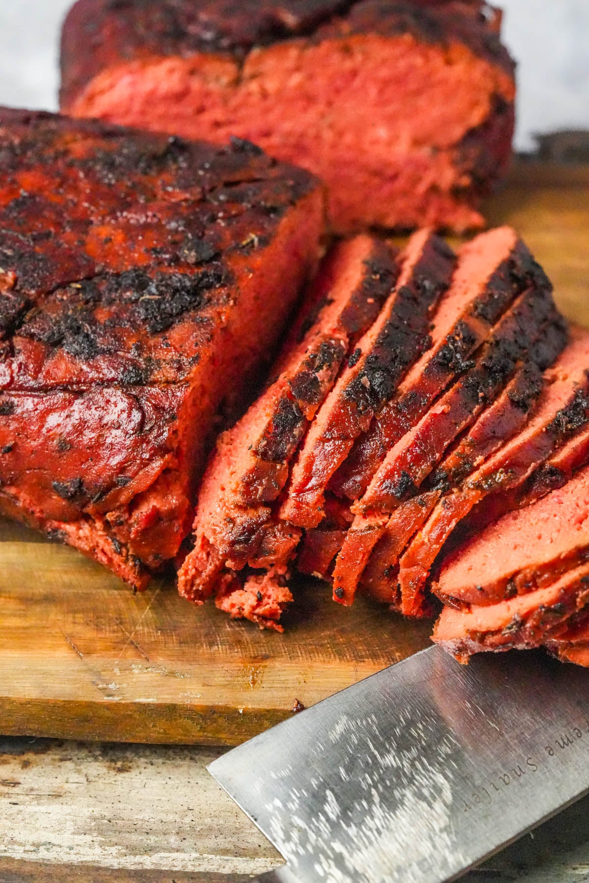 Sliced, smoked beet-red vegan roast beef on a wooden cutting board with a knife, showing a moist and smoky texture.