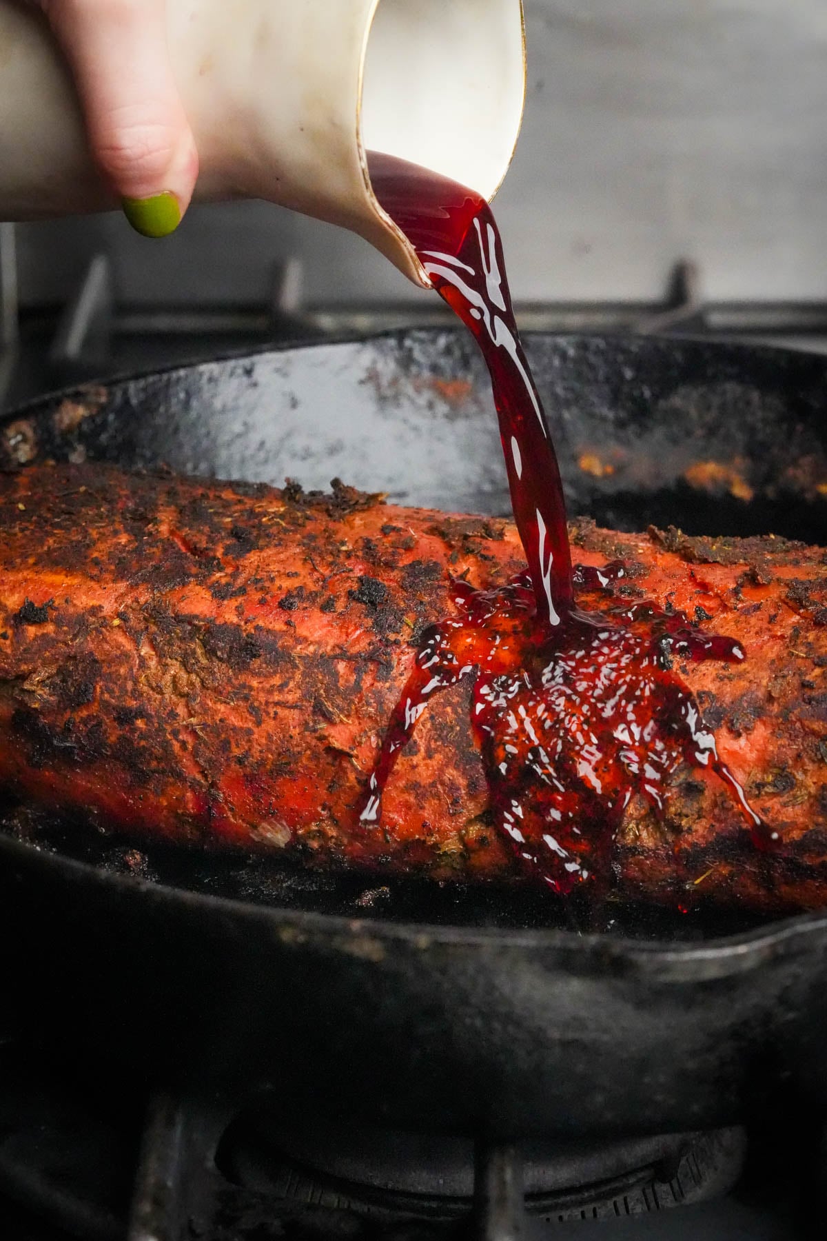 A hand pours red wine from a white pitcher onto a seasoned vegan roast beef in a black cast iron skillet on a stove.