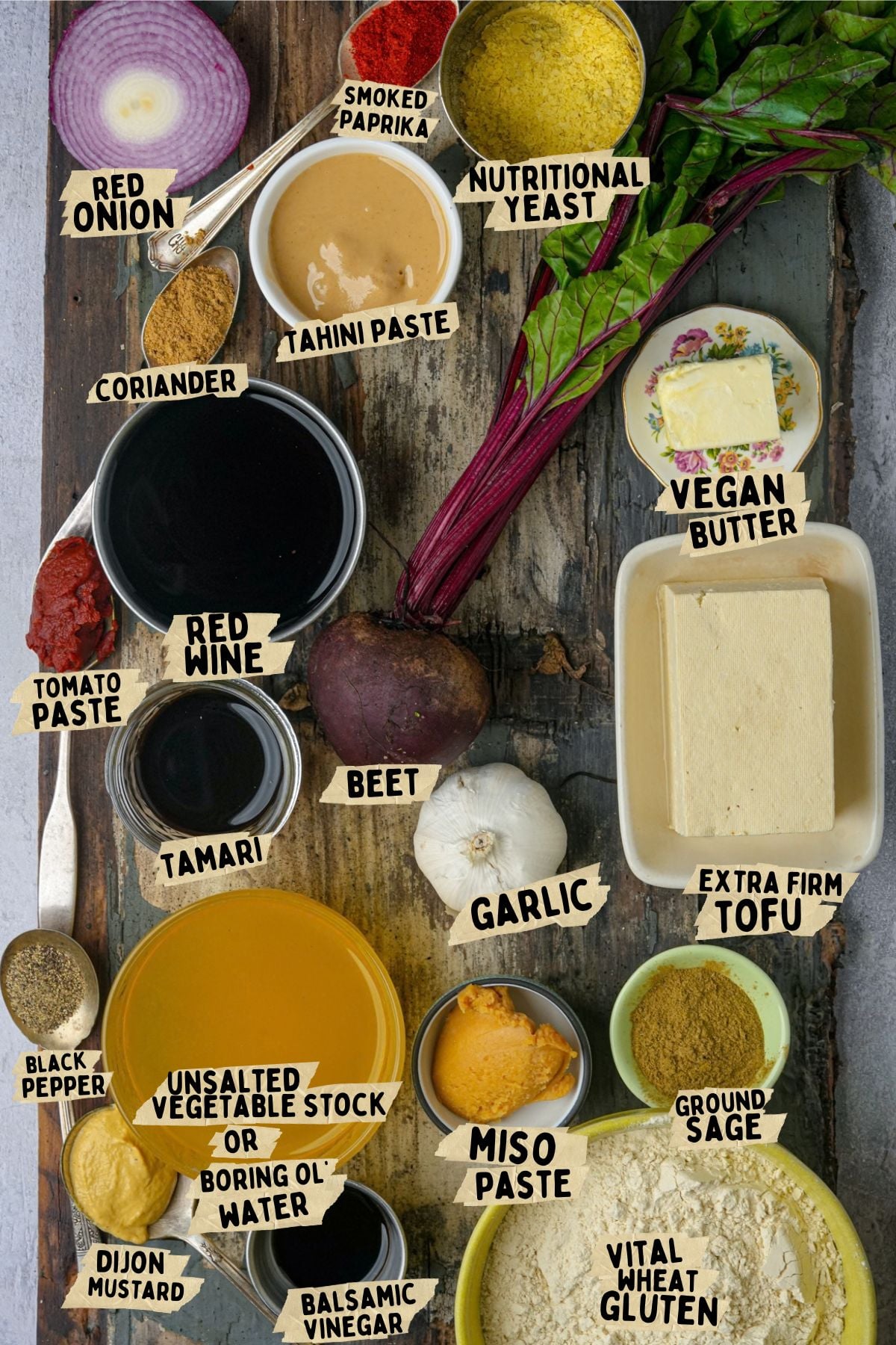 A tabletop with labeled ingredients for a vegan roast beef recipe, including beet, tofu, vegan butter, tahini, miso paste, vital wheat gluten, nutritional yeast, herbs, spices, vegetable stock, red onion, and garlic.